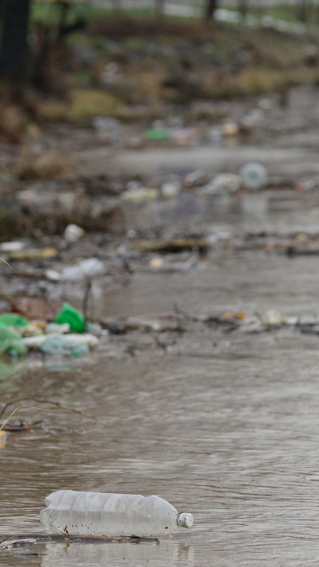 Dirty brown water flows with trash like plastic bottles in it.
